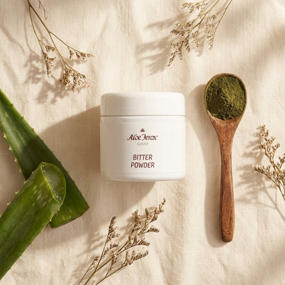 A container of Aloe Ferox Bitter Powder with a wooden spoon filled with green powder, surrounded by aloe ferox leaves and dried flowers on a light fabric background.