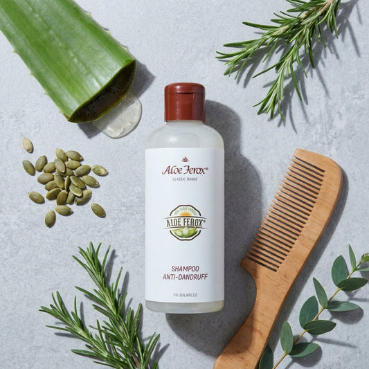 A bottle of Aloe Ferox shampoo with a wooden comb, green leaves, and seeds on a light gray background